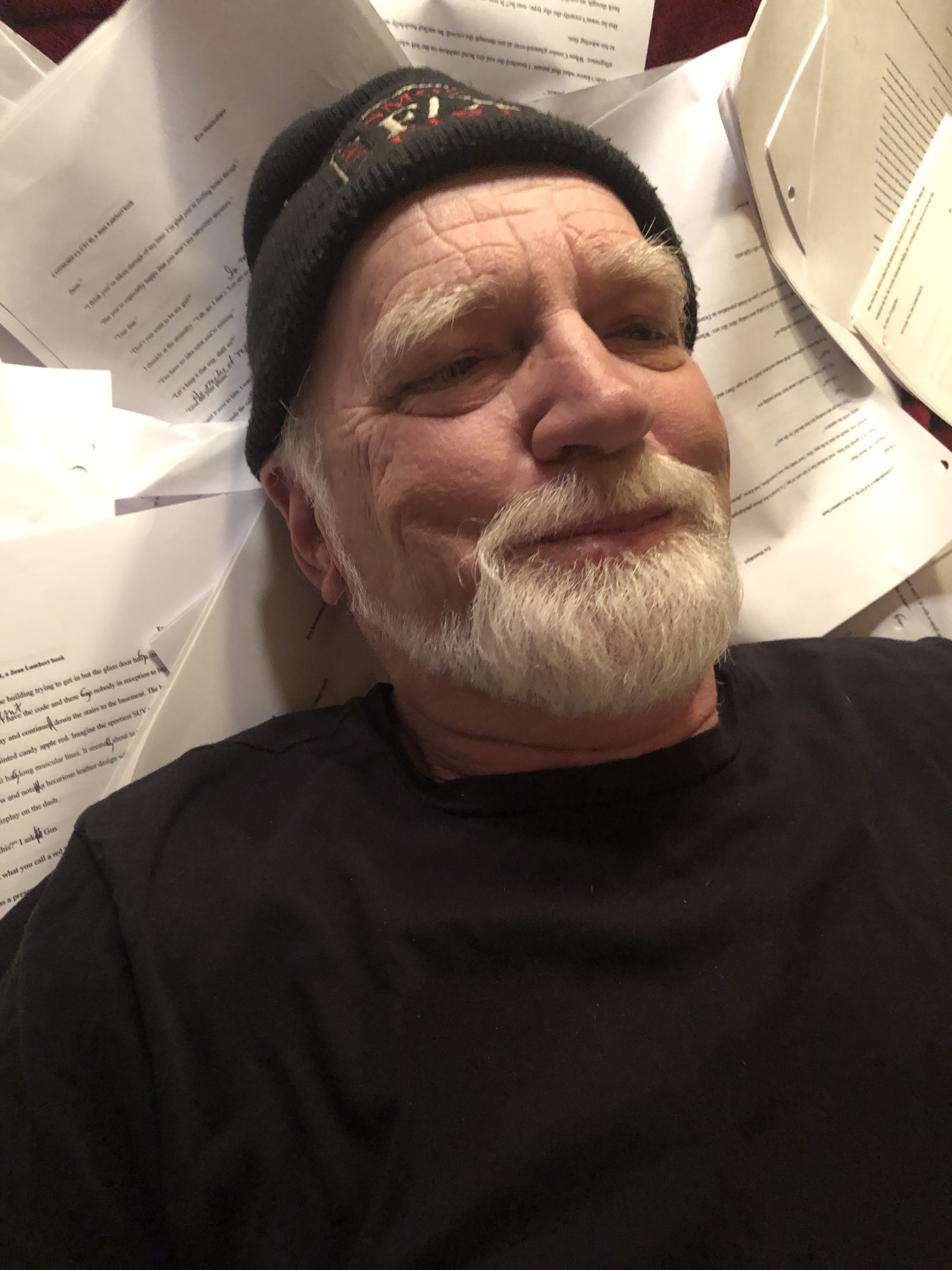 An older man with a white beard smiles while lying amidst scattered papers, suggesting deep engagement in writing or revision.