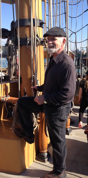 Man with a beard and cap stands on a ship's deck, resting one foot on a ship's rail. The scene captures a maritime environment with visible rigging and water in the background.