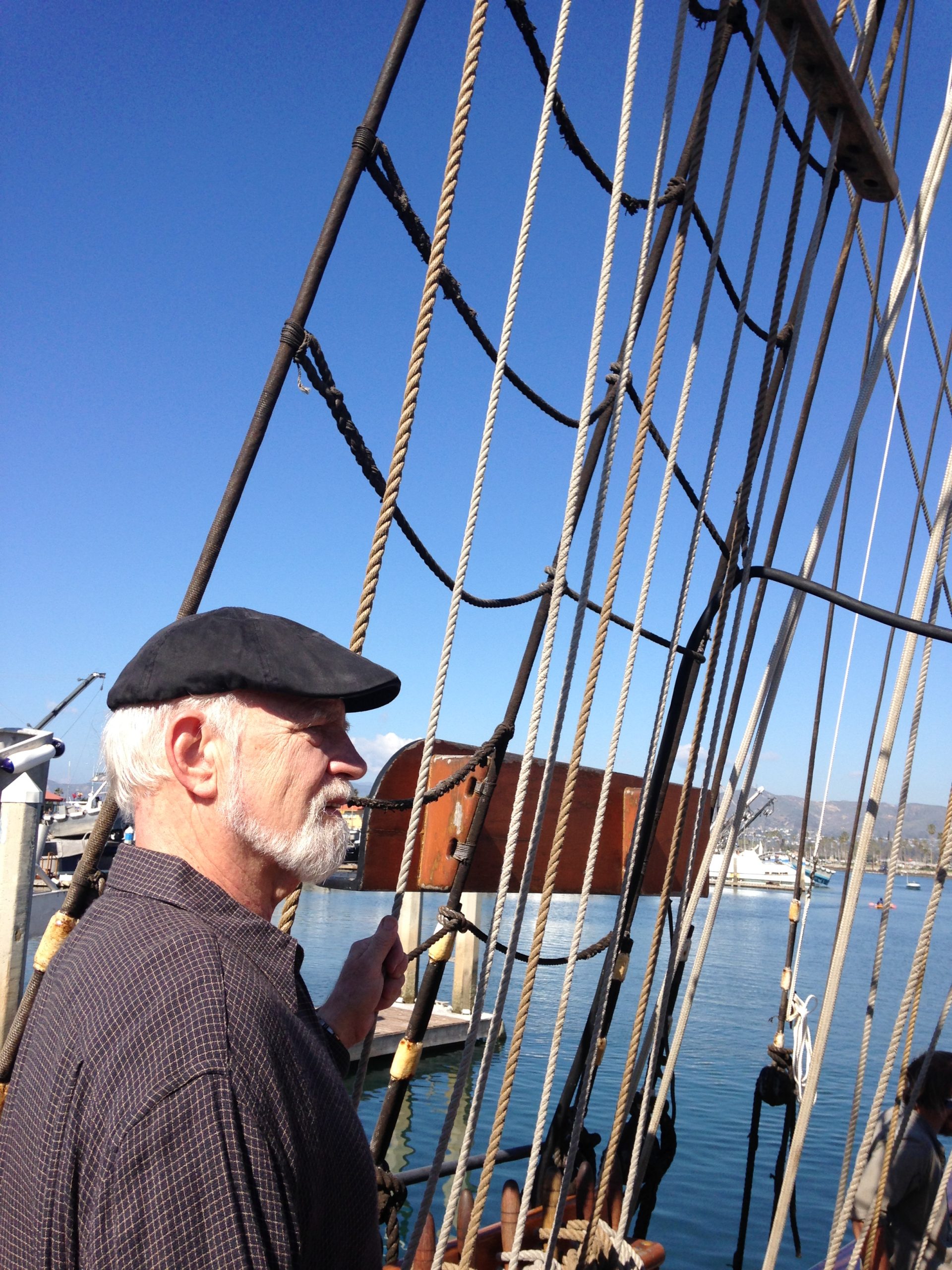 An older man with a beard stands aboard a ship, holding onto rigging against a clear blue sky, highlighting maritime life and craftsmanship.