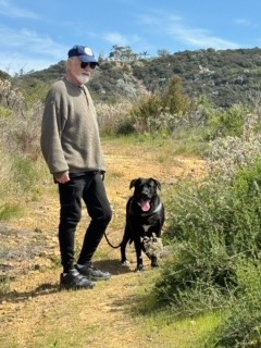 A man in a sweater and cap stands on a trail with a black dog, surrounded by greenery and hills under a blue sky. They appear to be enjoying a hike.
