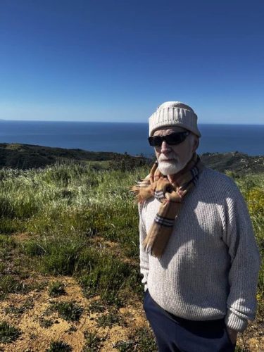 An elderly man wearing sunglasses, a knitted hat, and a scarf stands on a hillside overlooking the ocean under a clear blue sky.